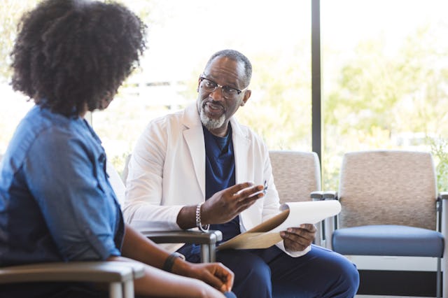 A doctor in a white coat is talking to a patient in a waiting area, holding a clipboard, with chairs...
