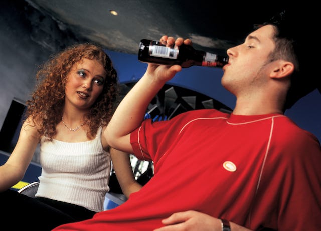 A young woman sits on a couch looking intently at a young man, who drinks from a bottle. The setting...