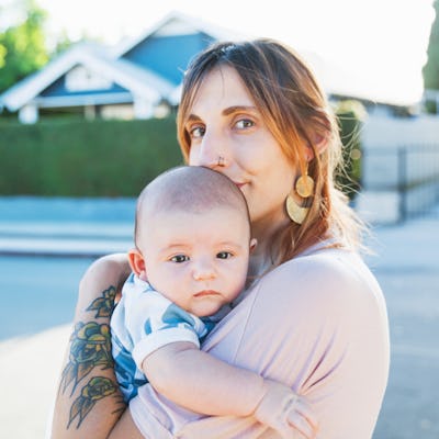 A mother with tattoos holding her newborn baby outdoors with a suburban house in the background