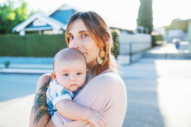 A mother with tattoos holding her newborn baby outdoors with a suburban house in the background