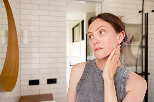 Mid adult woman examining her reflection in the bathroom mirror, touching her skin with a worried ex...
