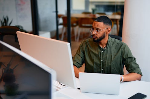Young adult programmer carefully looking at computer monitor while working on code in modern office,...