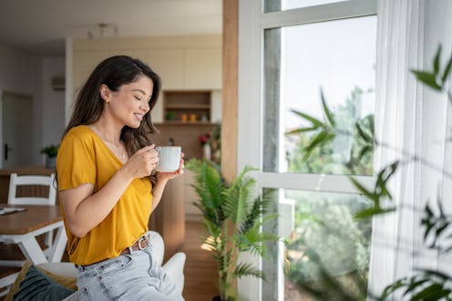 Serene young woman savoring a peaceful moment with a warm beverage, enjoying the bright view from he...
