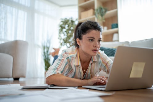 Young woman lying on the floor of her living room, working remotely on her laptop while reviewing va...
