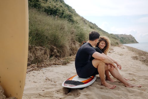 A couple in wetsuits sitting on a surfboard at the beach, engaging in a conversation. They are enjoy...
