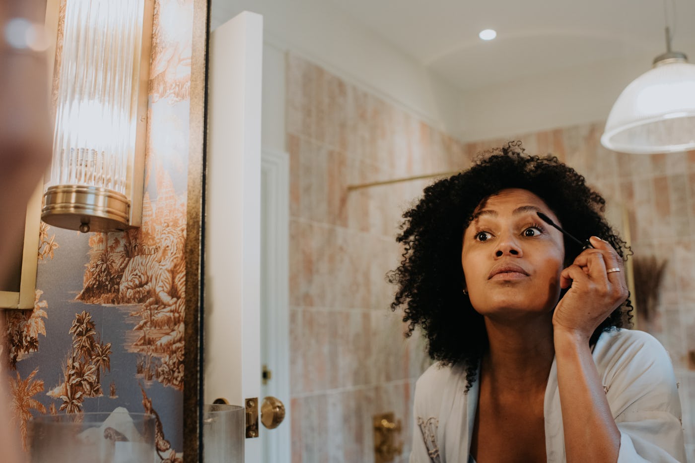 A beautiful young black woman concentrates as she applies mascara in a bathroom mirror.