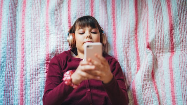 Teenage girl lies comfortably on a bed with a pink and white striped quilt, wearing large headphones...