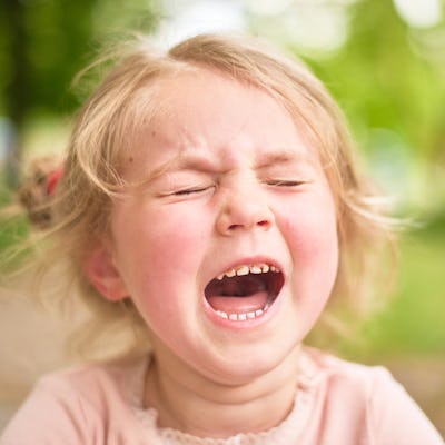 A young girl with light blonde hair is making an expressive face as she cries out, surrounded by greenery in a blurred background.
