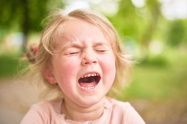 A young girl with light blonde hair is making an expressive face as she cries out, surrounded by gre...