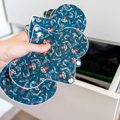 Hand holding reusable cloth menstrual pads near open cabinet drawer.