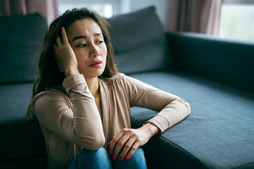 Asian woman is sitting on the floor and leaning with hand on sofa and looking through the window.