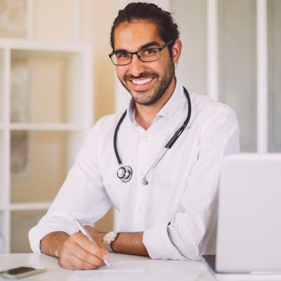 A smiling male doctor with glasses sits at a desk, wearing a white coat and stethoscope, writing notes with a laptop and phone nearby.