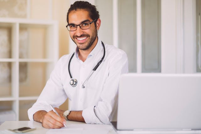 A smiling male doctor with glasses sits at a desk, wearing a white coat and stethoscope, writing not...