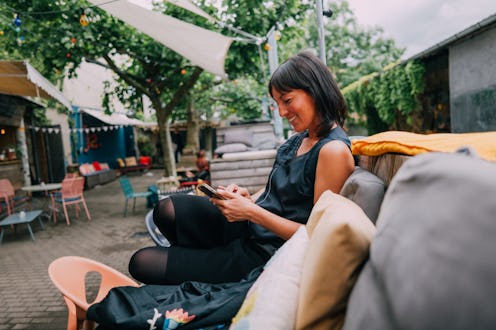 A cheerful woman enjoying outdoor leisure, sitting comfortably on a patio while browsing her smartph...