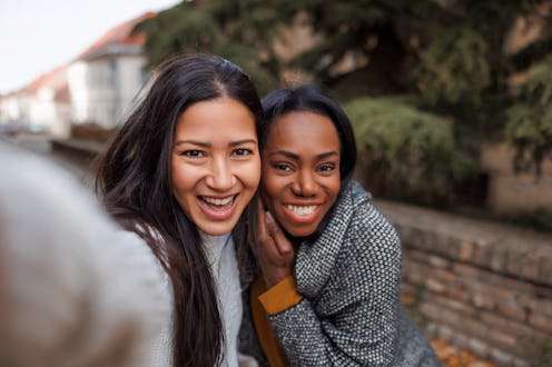 Female friends having fun taking selfies while walking down city streets, enjoying leisure time outd...