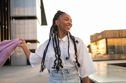 Young black woman with long braids smiles while holding a purple jacket, enjoying a sunny day in a m...