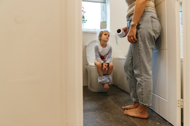 Young Caucasian girl sat on the toilet as her mum stands by the door holding the toilet roll and wat...