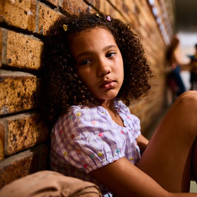 Schoolgirl sitting on floor with knees bent, leaning on wall, looking sideways with pensive expression in school corridor.
