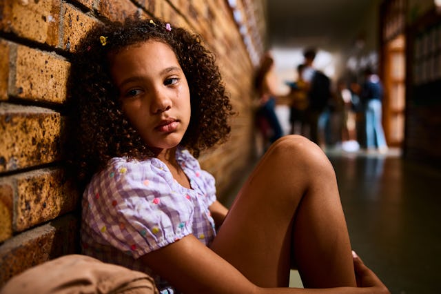 Schoolgirl sitting on floor with knees bent, leaning on wall, looking sideways with pensive expressi...