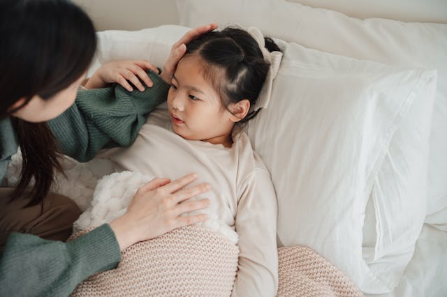 A caring adult gently tends to a young girl lying in bed, covered with a soft blanket, conveying com...