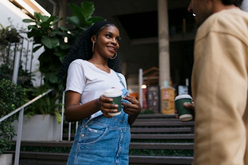 Happy young woman holds a coffee cup while engaging in conversation with a male friend outside. Dive...