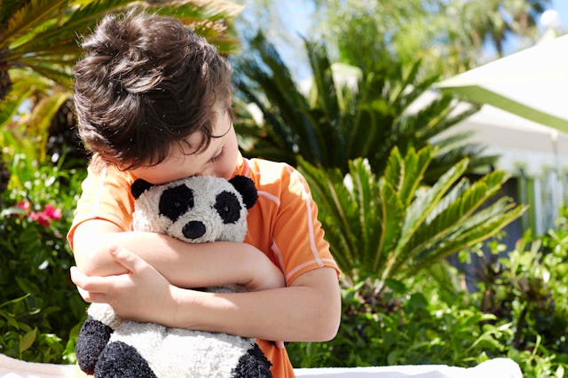 Waist up photo of a little boy standing on a lounge chair hugging his stuffed panda toy.He is wearin...
