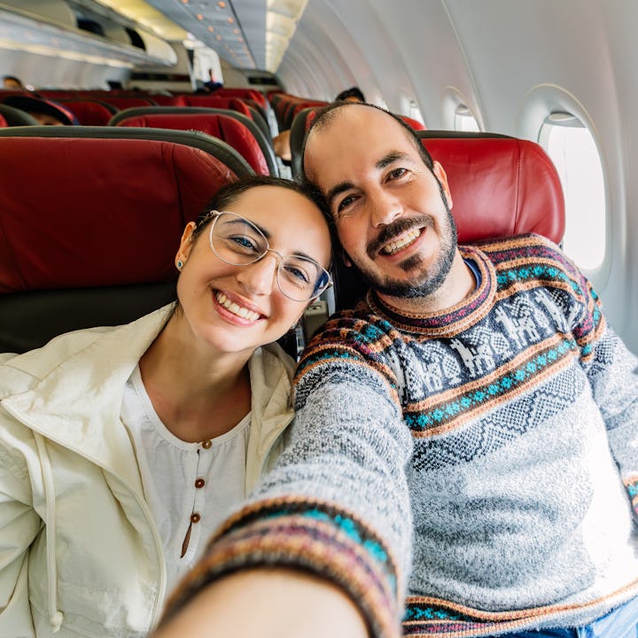 Smiling young adult couple taking selfie portrait inside the airplane before flying. Happy tourist e...