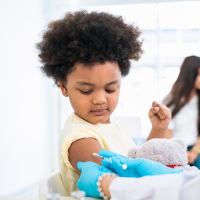 A young child with curly hair sits patiently as a healthcare professional administers a vaccine. Two other children are visible in the background.