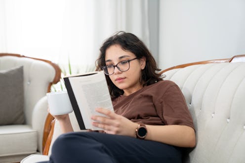 Photo of happy young woman reading book and drinking coffee at home relaxing on sofa