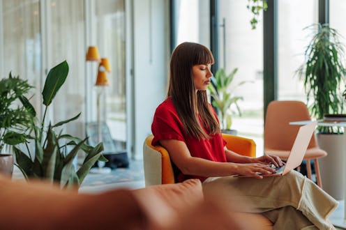 Young adult businesswoman concentrating on her work, typing on a laptop in a stylish and plant-fille...