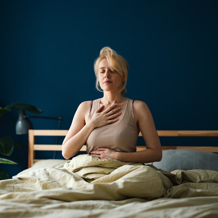 Happy woman in pyjamas sitting on a bed with closed eyes and doing breathing exercise at home.