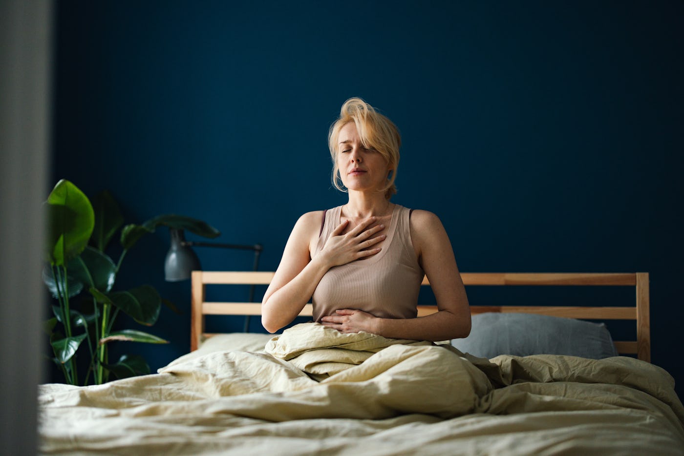 Happy woman in pyjamas sitting on a bed with closed eyes and doing breathing exercise at home.