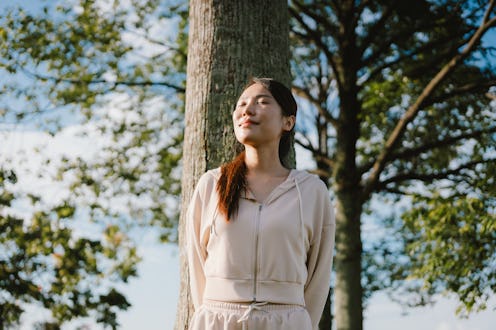 A young Asian woman stands calmly under a tree, eyes closed and basking in the warm sunlight, enjoyi...