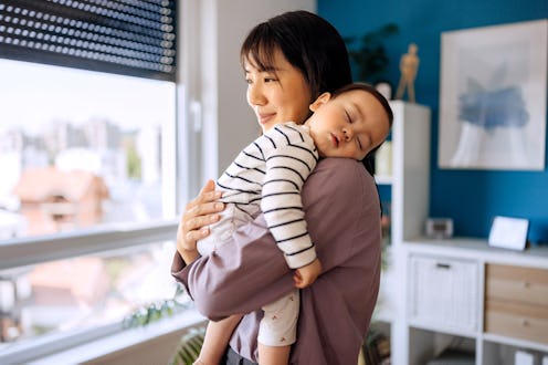 Young mom holding cute sleeping baby girl while standing next to a window in their home