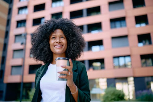 Young businesswoman with afro hair smiling while enjoying a takeaway coffee, gazing up at the impres...