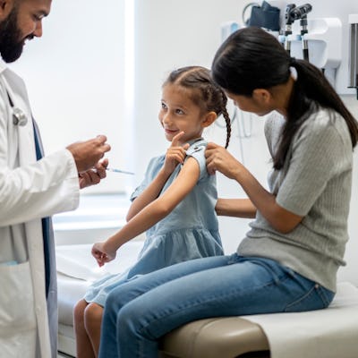 A young girl around 7 years old is at a doctor's office with her mother. They are both sitting on an examination table. She is looking at her doctor and pointing at her arm. The doctor is holding a syringe and ready to administer the flu shot onto his young patient's arm.
