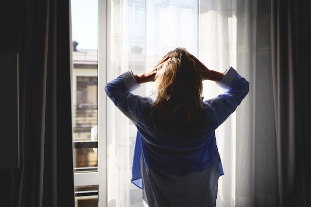 A woman standing by a window with her hands on her head, appearing contemplative or stressed. The in...