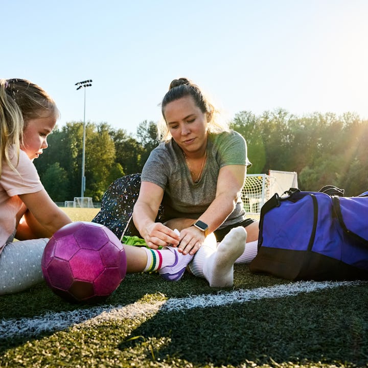 Young woman tying shoelaces of a girl sitting on a sports field and getting ready for soccer practic...