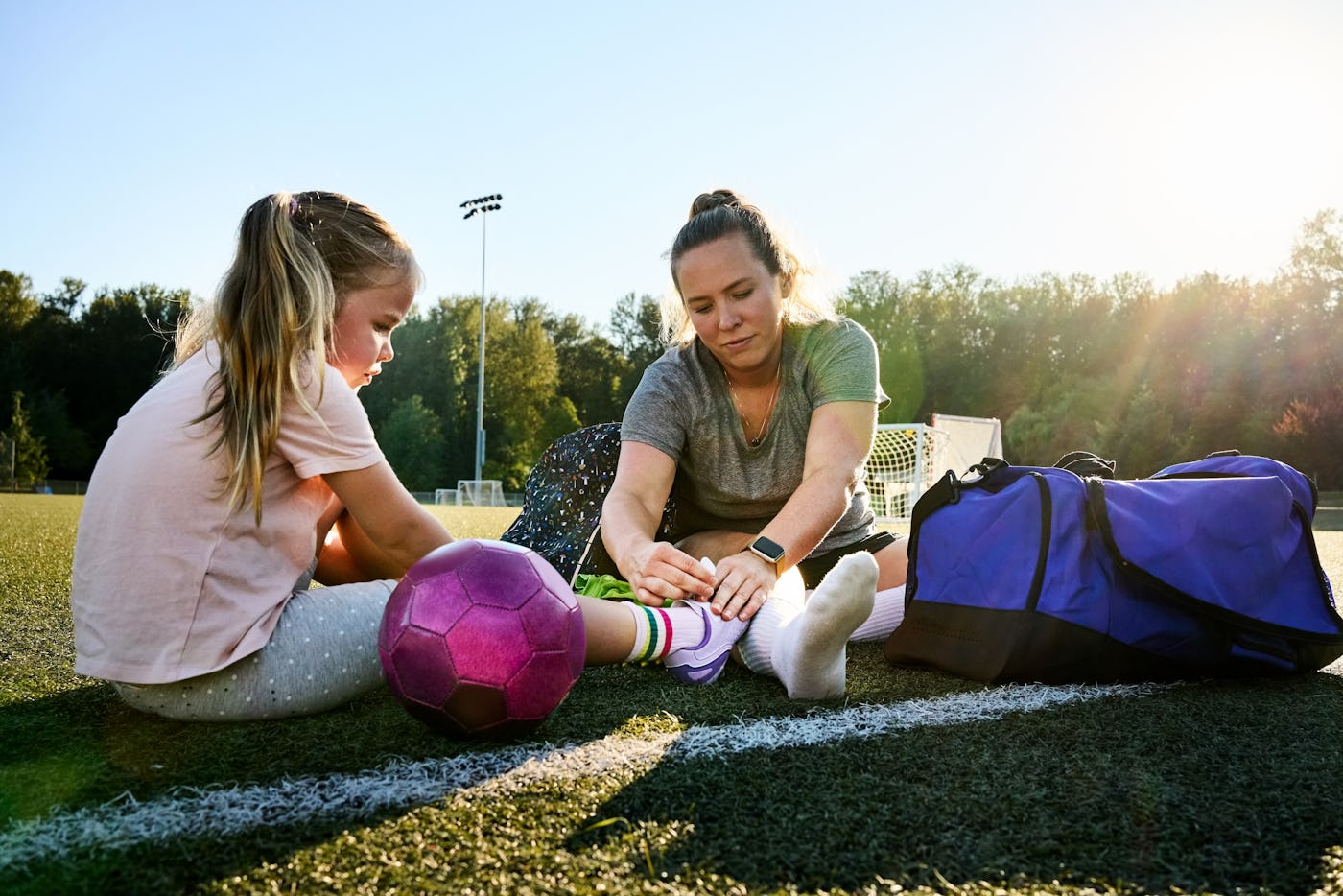 Young woman tying shoelaces of a girl sitting on a sports field and getting ready for soccer practice on summer day