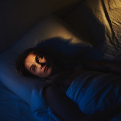 A woman lies awake in her bed, partially illuminated by dramatic lighting, evoking a mood of introspection or concern.