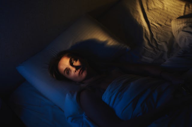 A woman lies awake in her bed, partially illuminated by dramatic lighting, evoking a mood of introsp...