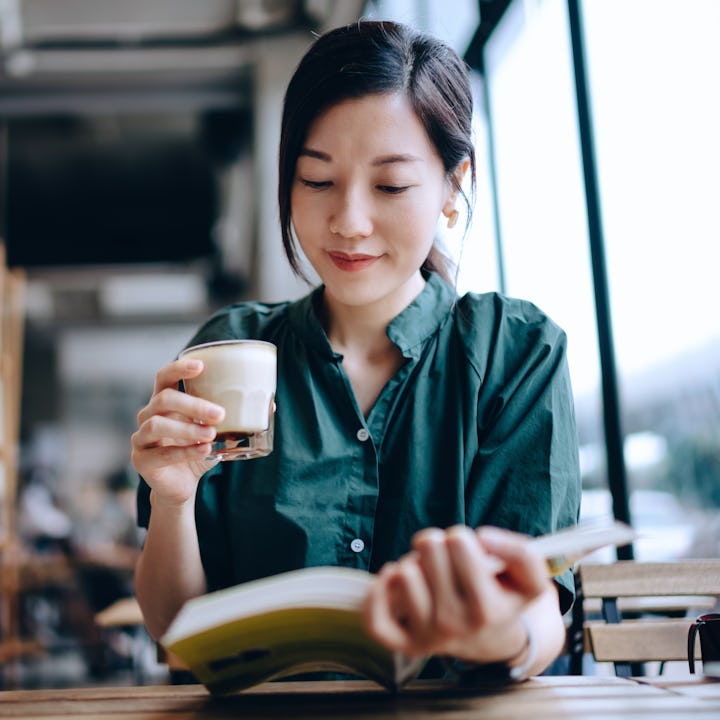 Young Asian woman taking a break, having a quiet time enjoying a cup of coffee and reading book in c...