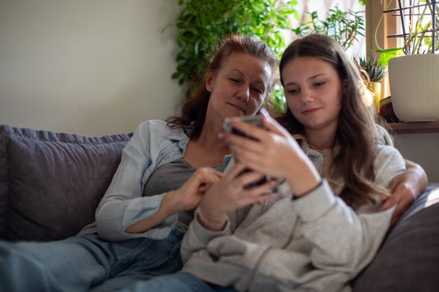 Mother and daughter are sitting on the sofa in the living room, watching something on their phone, a...