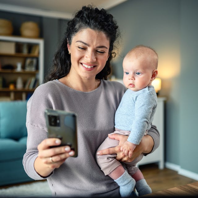 A smiling mother holds her baby while looking at a smartphone. They are in a cozy living room with s...