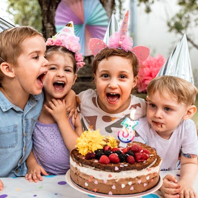 Cute little girl celebrating 3rd birthday with friends in backyard, blowing birthday cake candles.