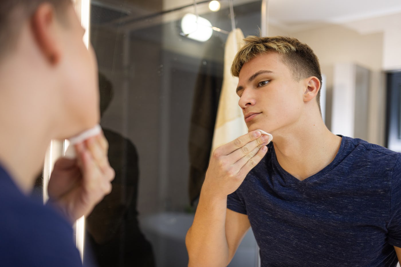 A young man is carefully grooming himself in front of a bathroom mirror, reflecting a routine of personal care and self-awareness.