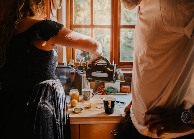 A couple in a kitchen prepare a hot drink. The pregnant woman pours some boiling water into a cup fr...