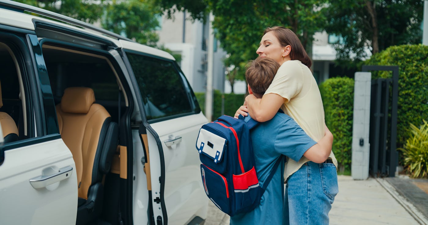 Young mother says goodbye to her son at the car door, giving him a final hug before he leaves for his first day of school, capturing a moment of care and excitement. Back to school concept.