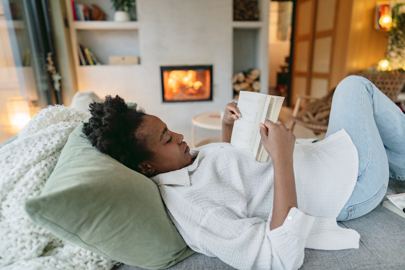 Photo of a young African American woman reading a book in her living room by the fireplace