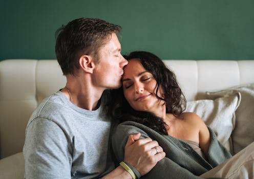 A smiling man and woman, a loving couple, sit close together in their comfortable bed, sharing a qui...
